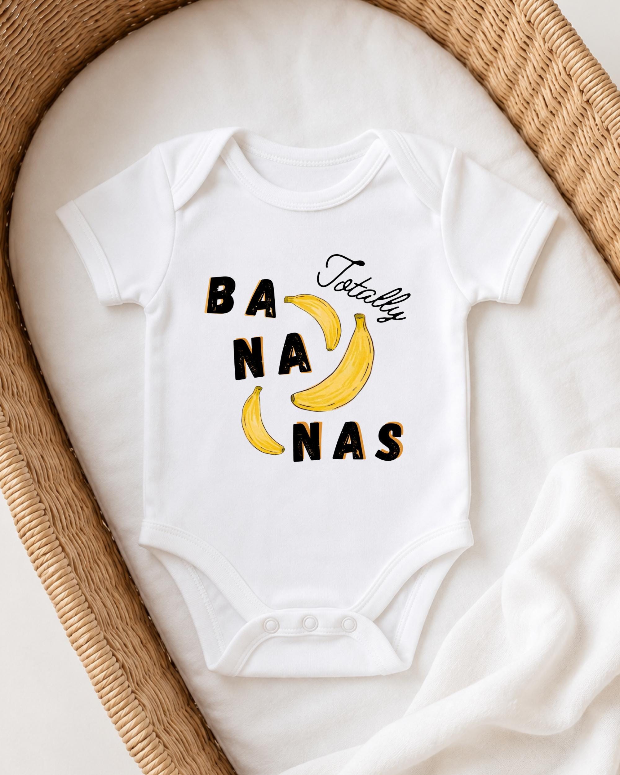 A white onesie with the text "Bananas Totally" printed on it, placed on a white blanket inside a woven basket.