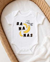 A white onesie with the text "Bananas Totally" printed on it, placed on a white blanket inside a woven basket.