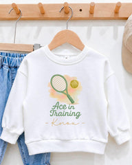 A white sweatshirt with a tennis racket and ball graphic printed on it, along with the text "Ace in Training" and "Knox" printed below.