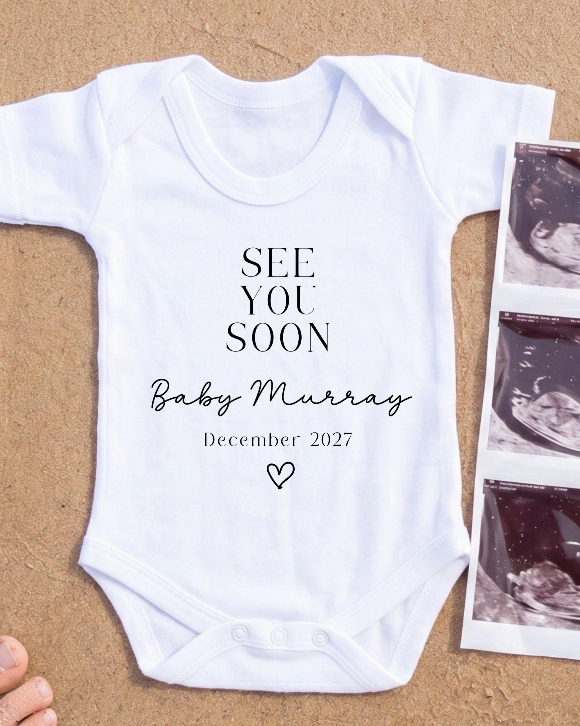 A white onesie with the text "See you soon Baby Murray December 2027" printed on it, along with a heart symbol. The onesie is placed next to a photograph of an ultrasound image.