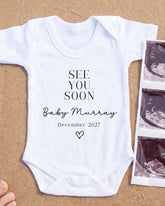 A white onesie with the text "See you soon Baby Murray December 2027" printed on it, along with a heart symbol. The onesie is placed next to a photograph of an ultrasound image.