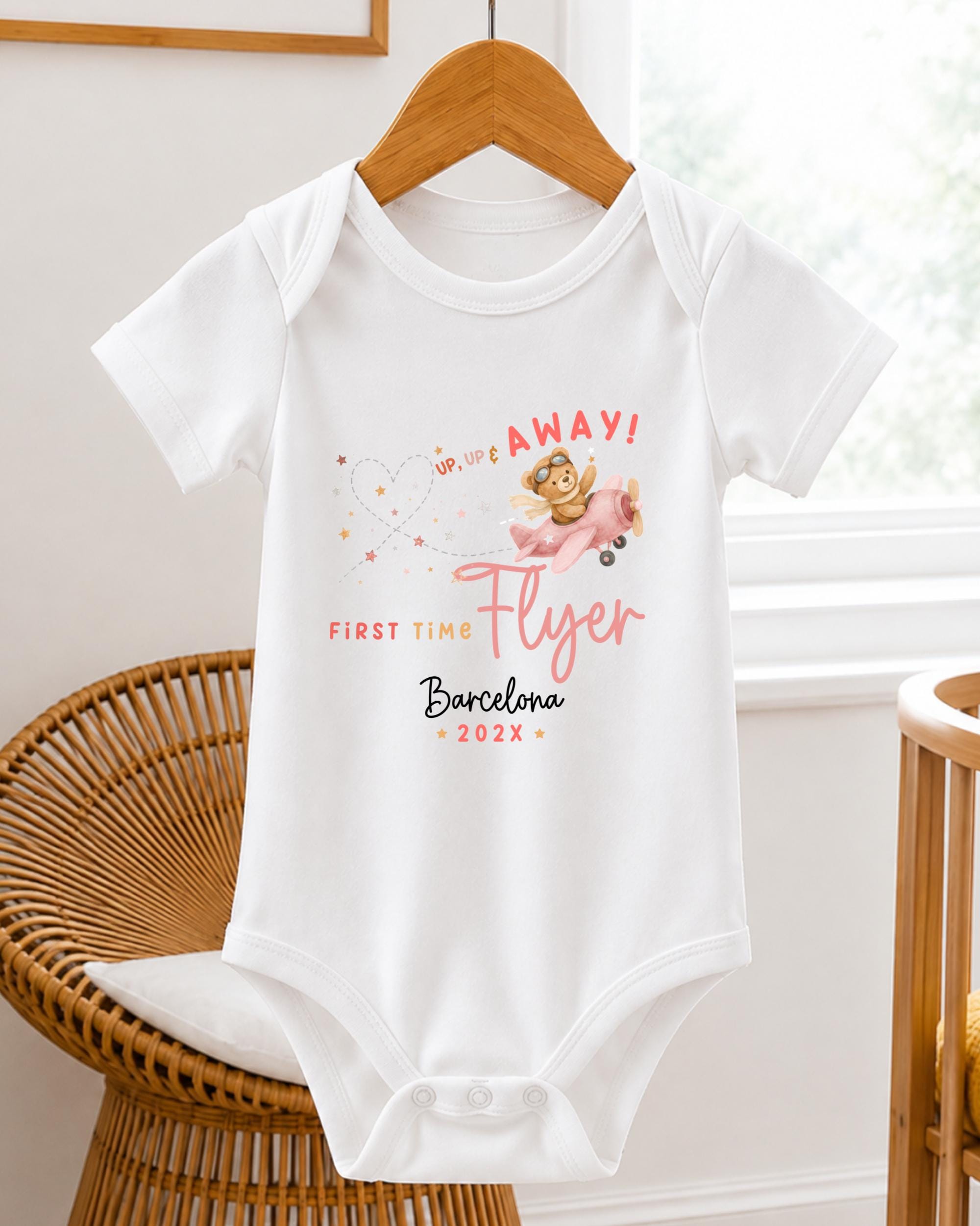 A white baby onesie with a pink and yellow design, featuring a cartoon character and text that says "UP! UP! UP! Away! First time flyer Barcelona 20XX".