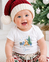 A smiling baby wearing a red and white knit hat and a white t-shirt with a Christmas-themed design, sitting in front of a Christmas tree.
