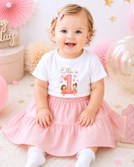 A smiling baby girl wearing a white t-shirt with a pink skirt and a pink number 1 sign, sitting on a pink and white background with pink and gold decorations.