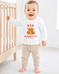 A smiling baby boy wearing a white sweatshirt with a pancake and breakfast design, standing next to a wooden crib.