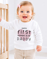 A smiling baby girl wearing a white sweatshirt with text that says "Happy first birthday as my Daddy".