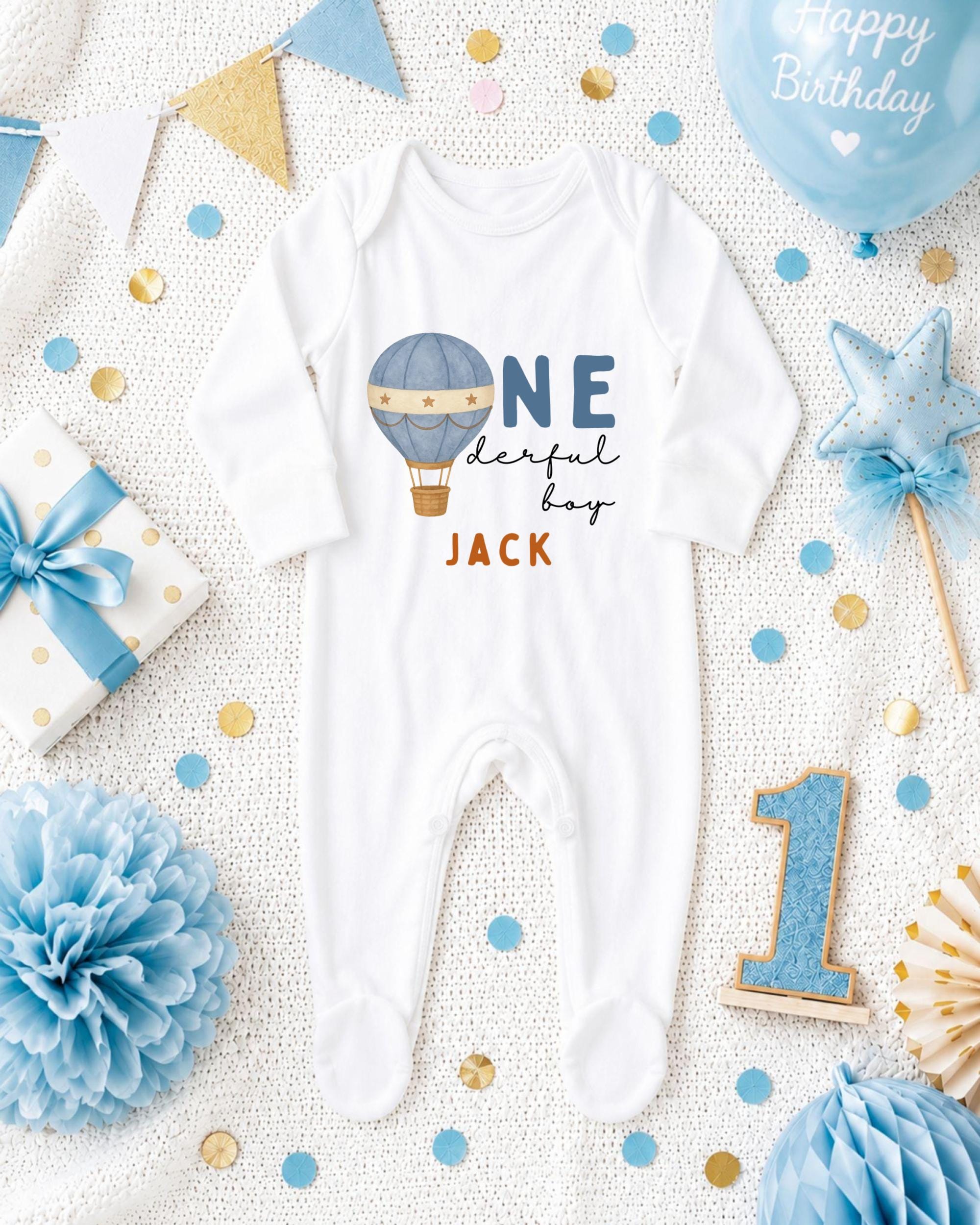 A white onesie with a hot air balloon design and the text "ONE derful boy JACK" printed on it, surrounded by birthday decorations such as a blue balloon, a star, and confetti.