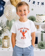 A young boy wearing a white t-shirt with a lobster graphic and the text "CLAW SOME BOY" is smiling at the camera. The background includes balloons, a cake, and other party decorations.