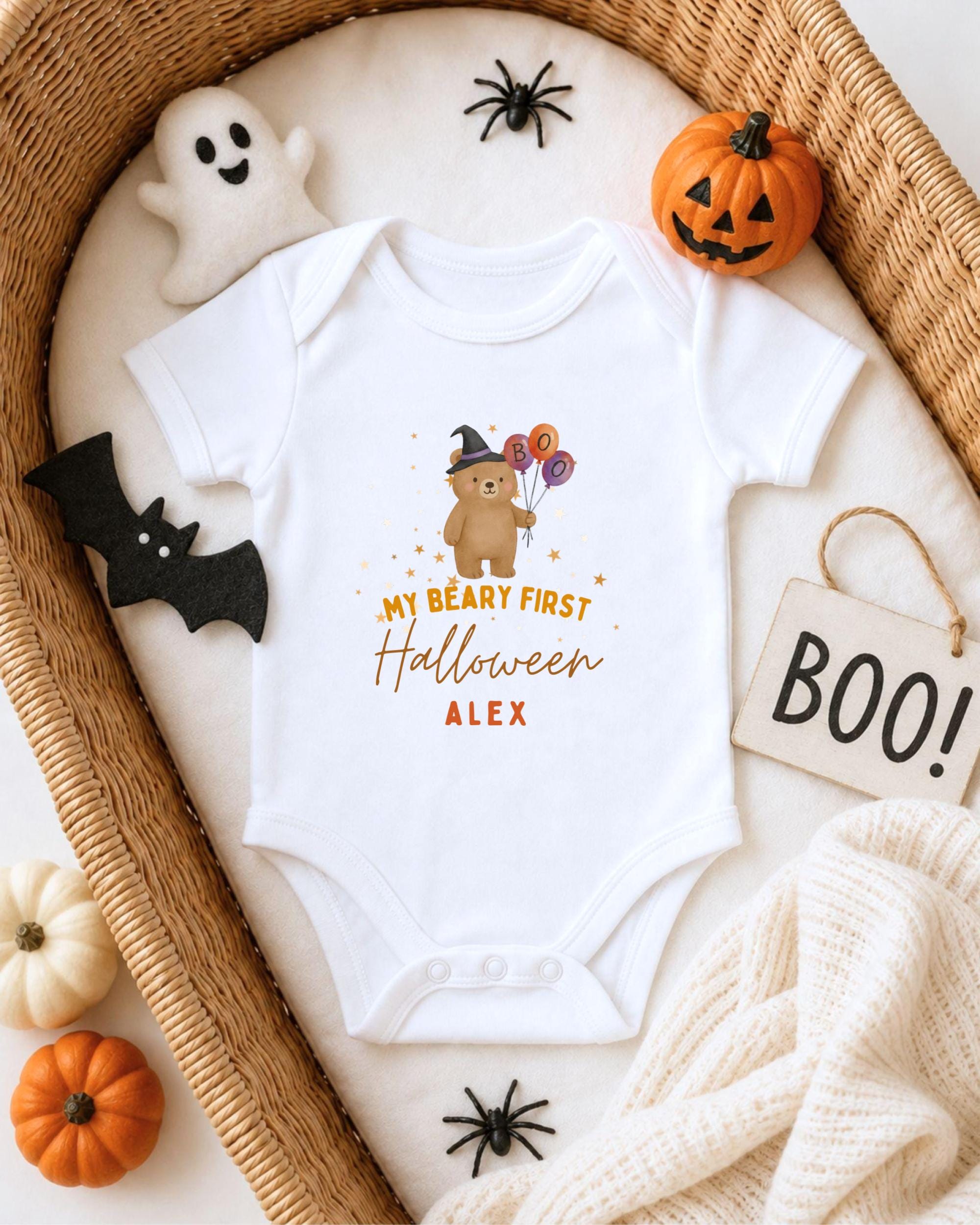 A white onesie with a cartoon bear and Halloween-themed text, surrounded by Halloween decorations such as a ghost, pumpkin, and a sign that says "BOO!".