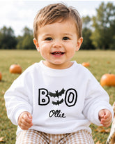 A smiling baby boy wearing a white sweater with the text "BOO OLLIE" printed on it, sitting in a field with pumpkins in the background.