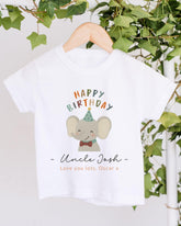 A white t-shirt with a colorful graphic of an elephant wearing a party hat and the text "Happy Birthday Uncle Josh" printed on it, hanging on a wooden hanger.