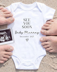 a white onesie with the text "See you soon Baby Murray" and the date "December 2027" printed on it, surrounded by sand and a black and white photograph of a baby's belly.