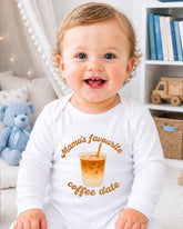A smiling baby wearing a white onesie with a coffee cup graphic and text, sitting in a room with a teddy bear and bookshelf in the background.