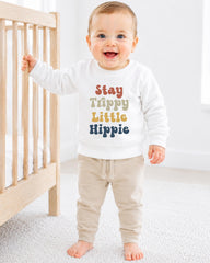 A smiling baby boy wearing a white sweatshirt with the text "Stay Trippy Little Hippie" printed on it, standing next to a wooden crib.