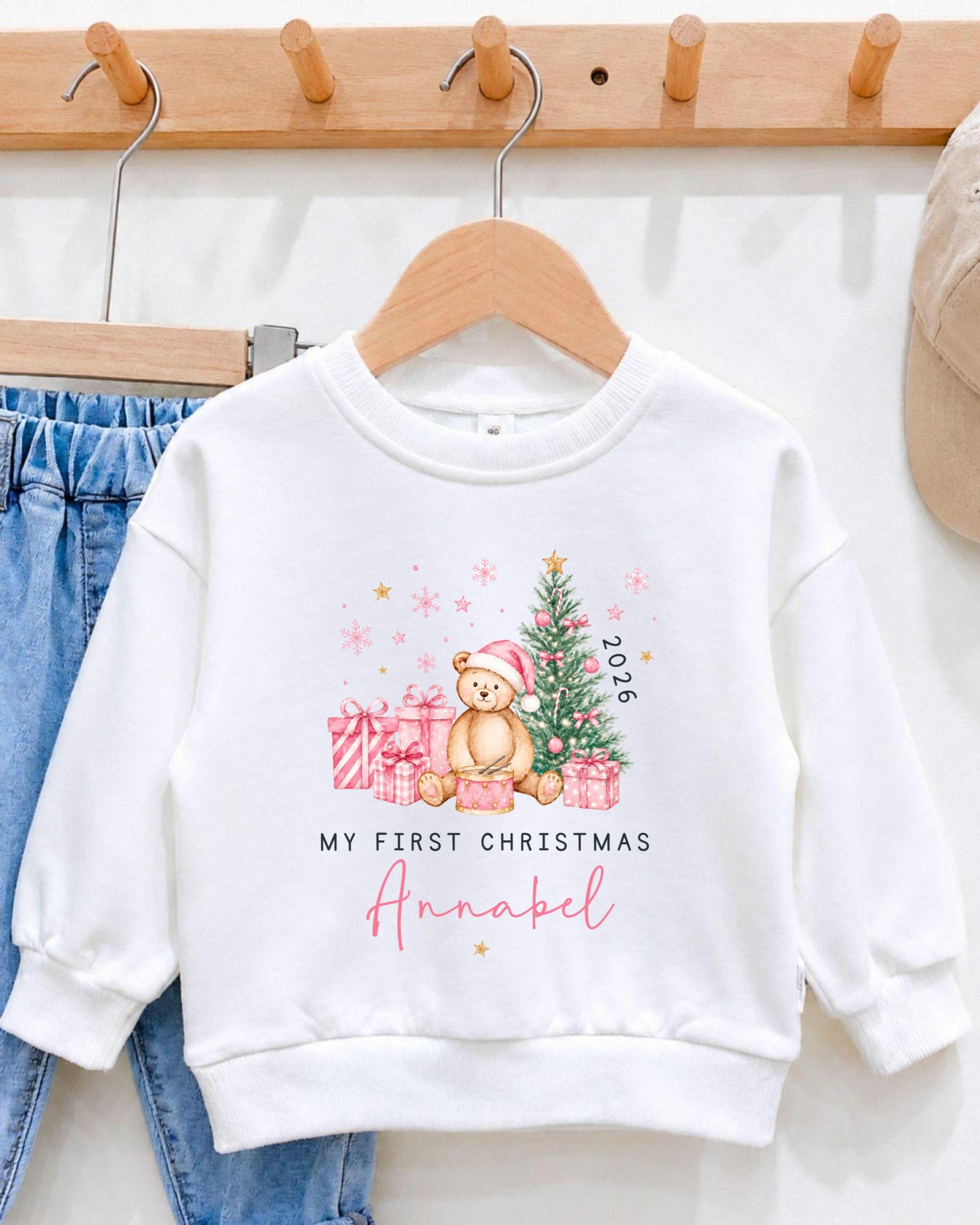 A white sweater with a Christmas-themed design, including a teddy bear, presents, and a tree, hanging on a wooden hanger.