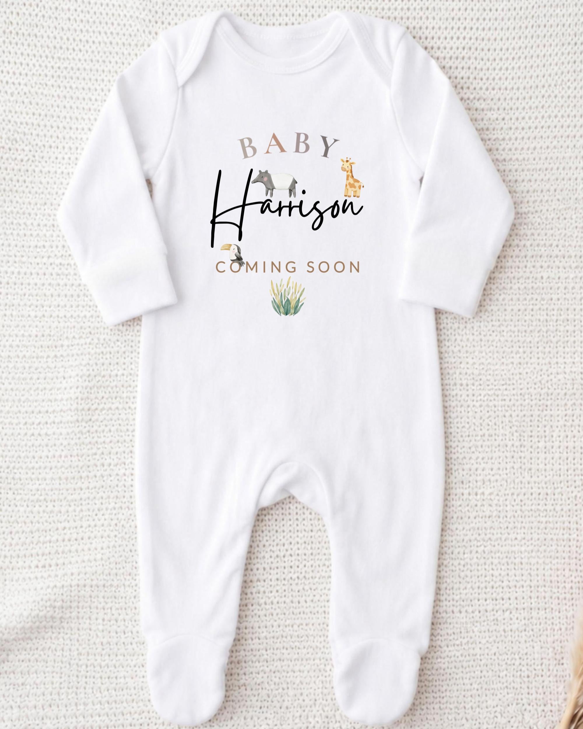 A white onesie with the text "BABY Harrison COMING SOON" printed on it, along with a small illustration of a giraffe and a plant.
