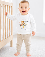 A smiling baby boy wearing a white sweatshirt with a cartoon character and the name "Ashley" printed on it, standing in front of a wooden crib.