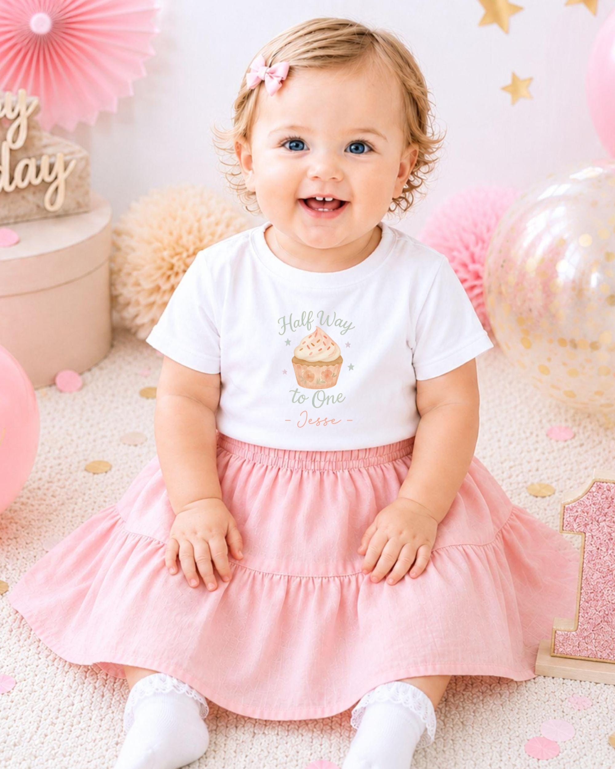 A smiling baby girl wearing a white t-shirt with a cupcake design and a pink skirt, sitting on a pink and white background with balloons and confetti.