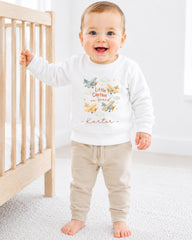 A smiling baby boy wearing a white sweatshirt with a graphic of airplanes and the text "Little Captain on brand" is standing next to a wooden crib.