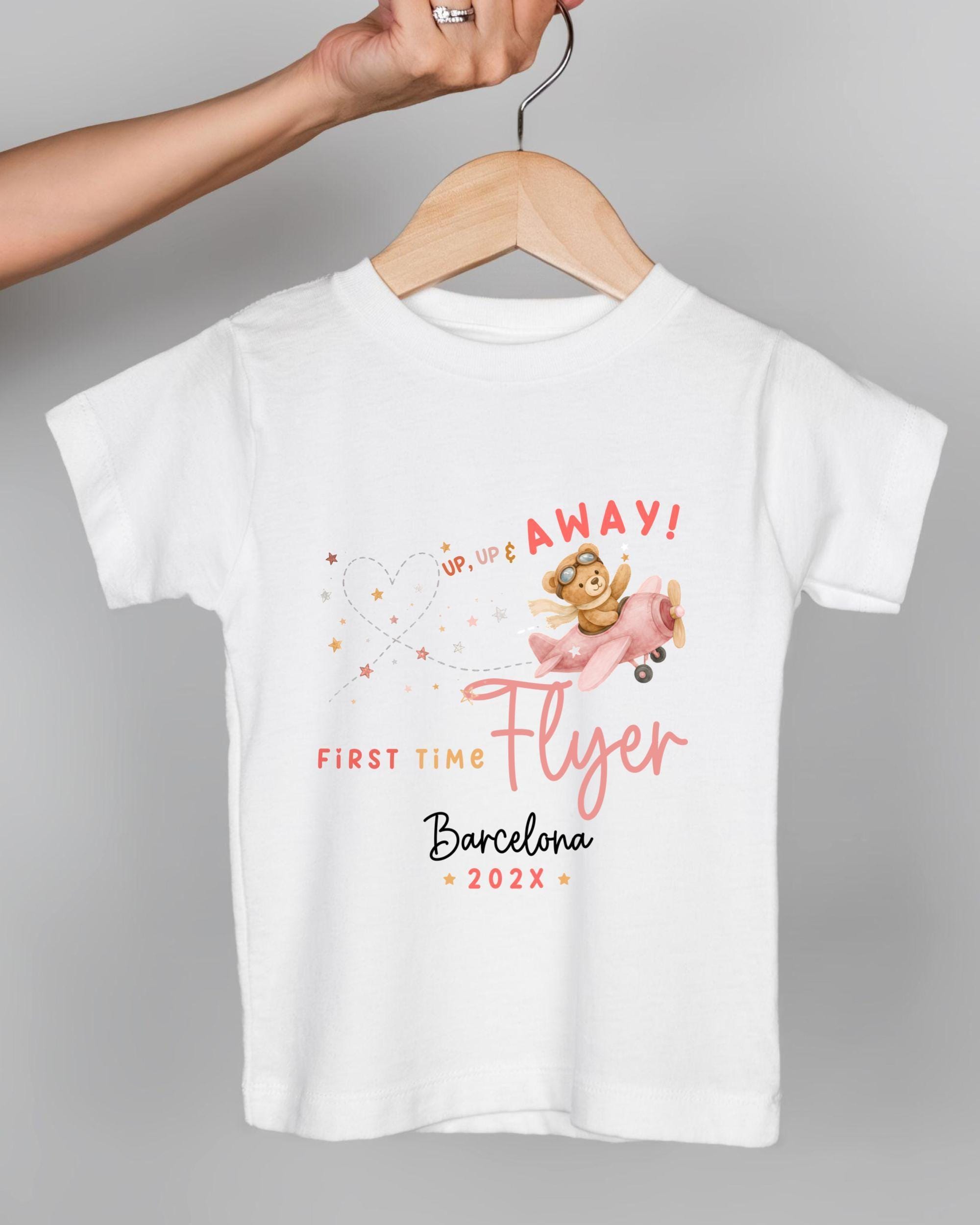 A white t-shirt with a graphic design featuring a cartoon character flying in a plane and the text "UP UP E AWAY! FIRST TIME FLYER Barcelona 20x" printed on it.