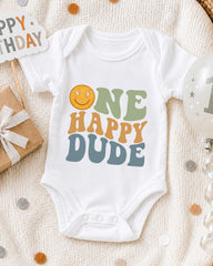 A white onesie with the text "ONE HAPPY DUDE" printed on it, surrounded by birthday decorations and a gift box.