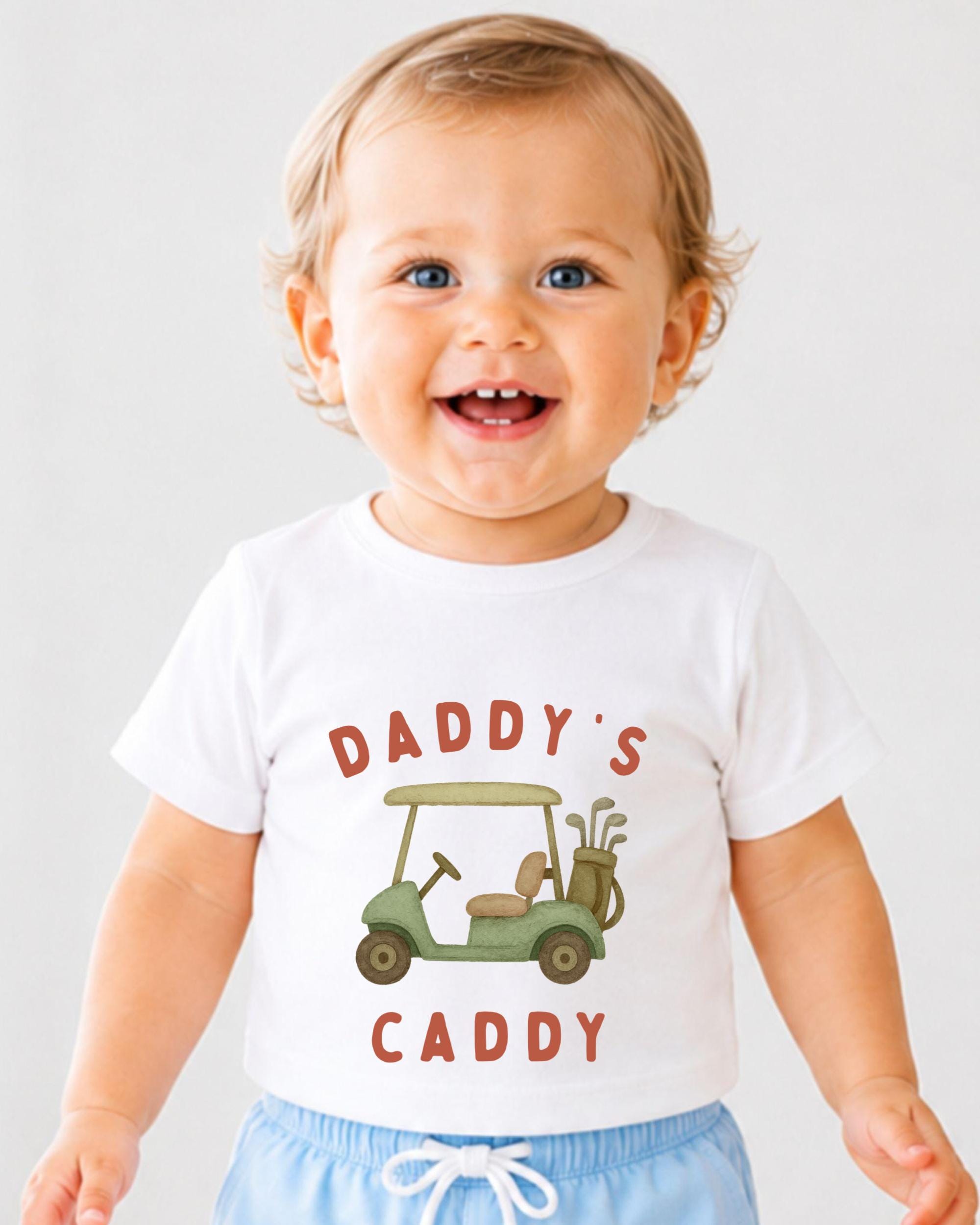 A smiling baby wearing a white t-shirt with a golf cart graphic and the text "DADDY'S CADDY" printed on it.