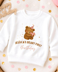 A white sweatshirt with a teddy bear and a birthday hat printed on it, along with the text " Jessica's Beary First Birthday".
