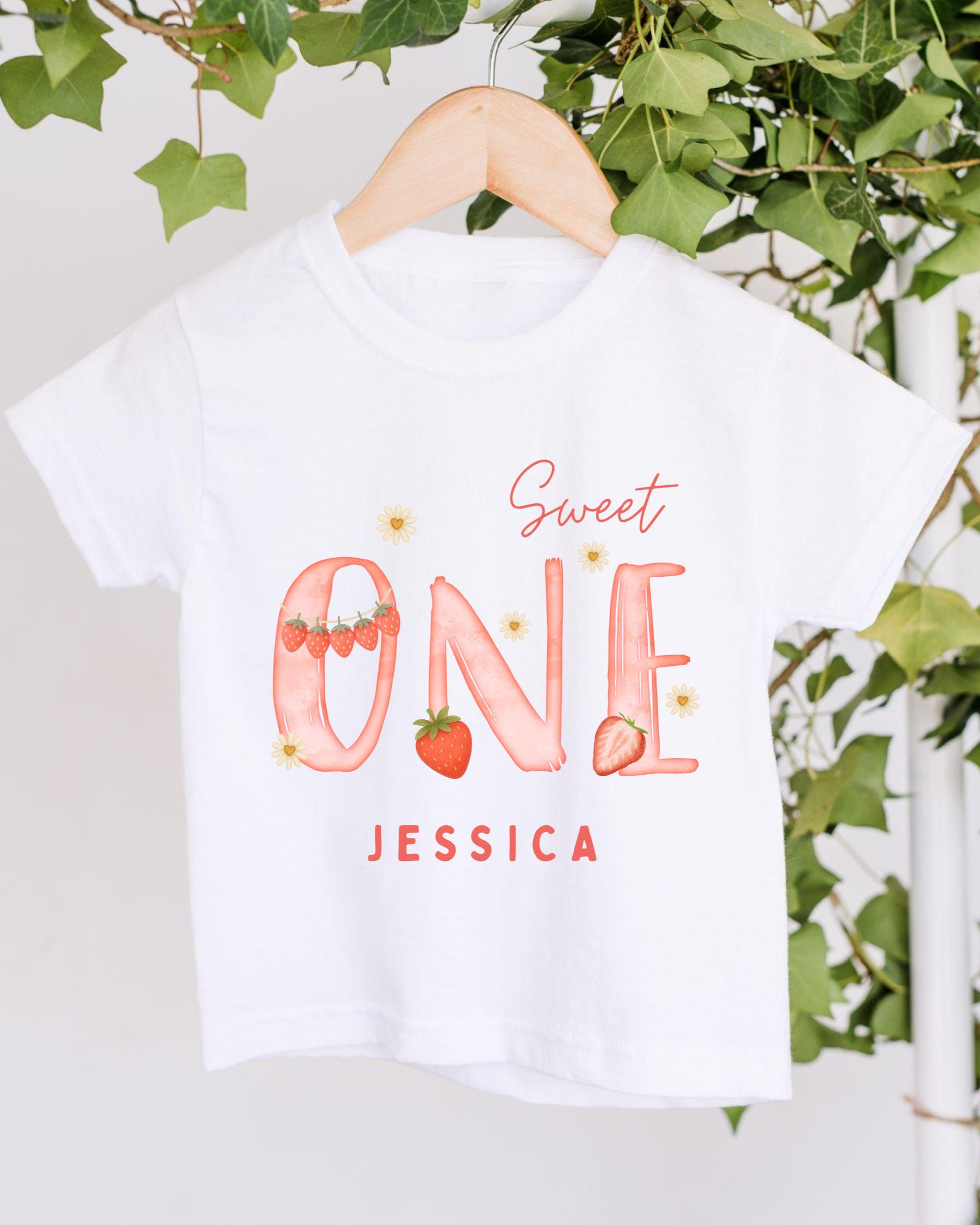 A white t-shirt with a personalized text and design printed on it, hanging on a wooden hanger.
