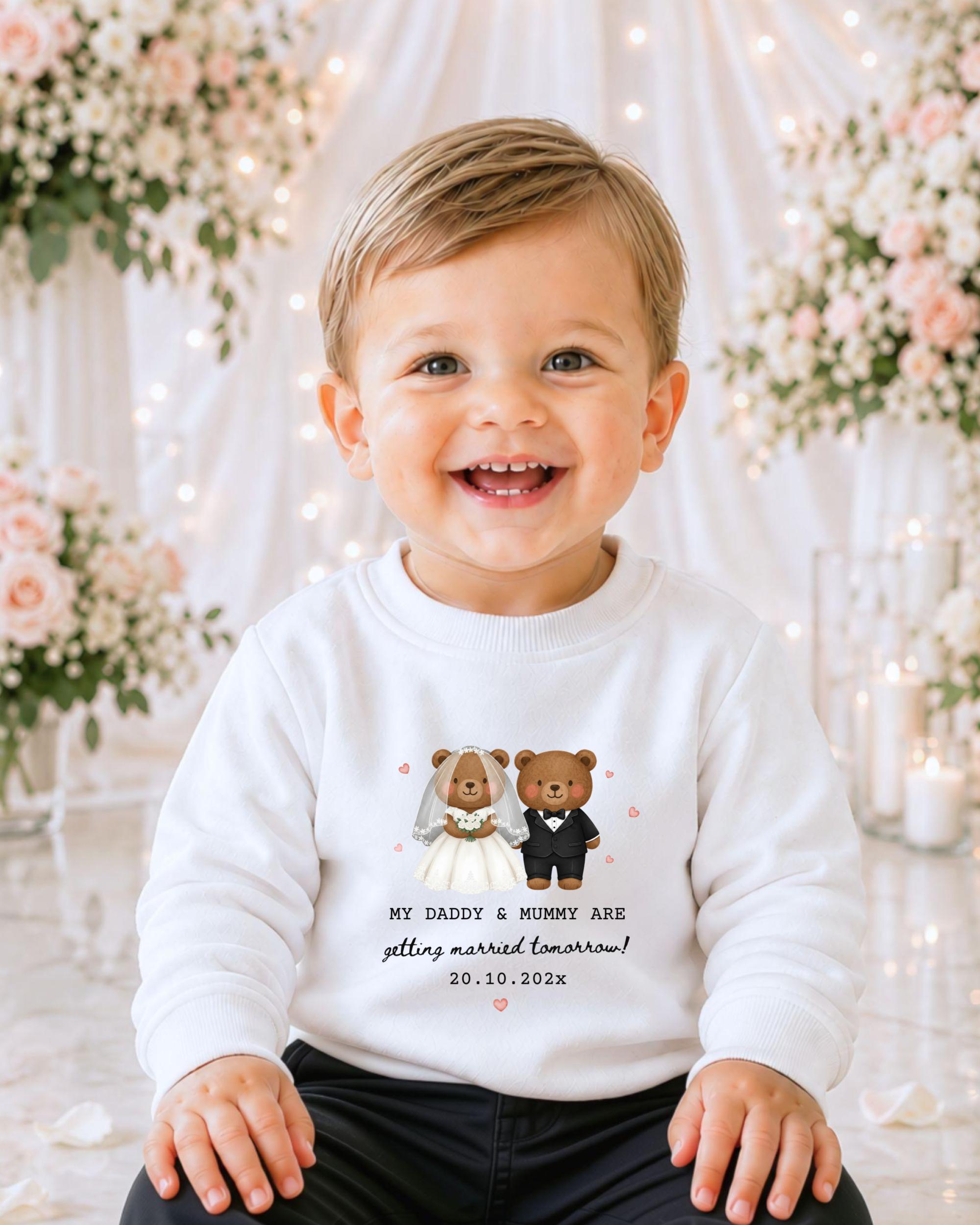 A smiling baby boy wearing a white sweater with a graphic of two bears dressed as a bride and groom, standing in front of a floral backdrop.