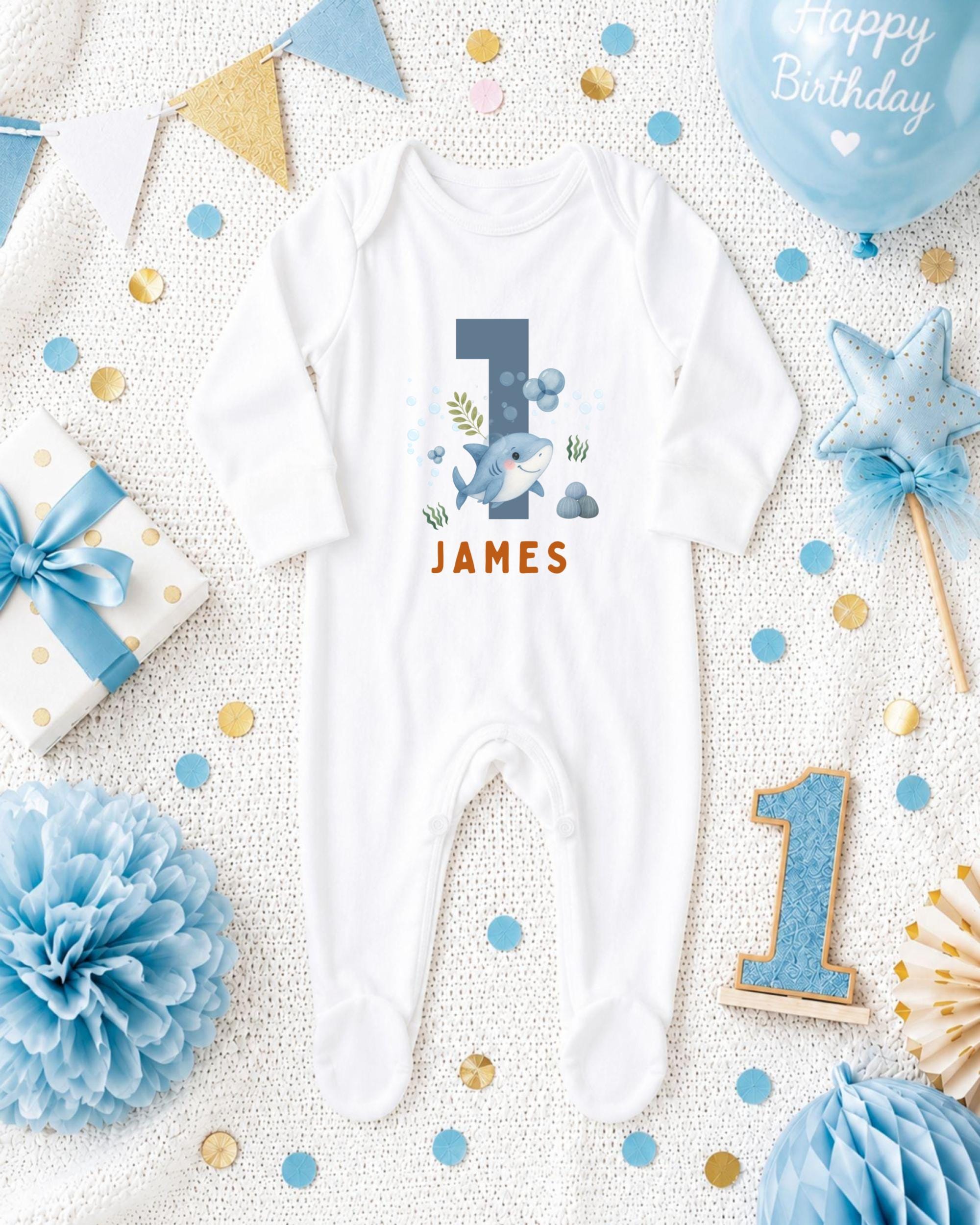 A white onesie with a shark design and the name "James" printed on it, surrounded by blue and gold decorations, confetti, and a blue balloon with the text "Happy Birthday".