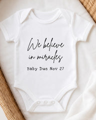 A white baby onesie with the text "We believe in miracles" printed on it, along with the date "Baby Due Nov 27".