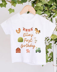 A white t-shirt with a graphic design featuring farm animals and a banner that says "Ellie's First Birthday".