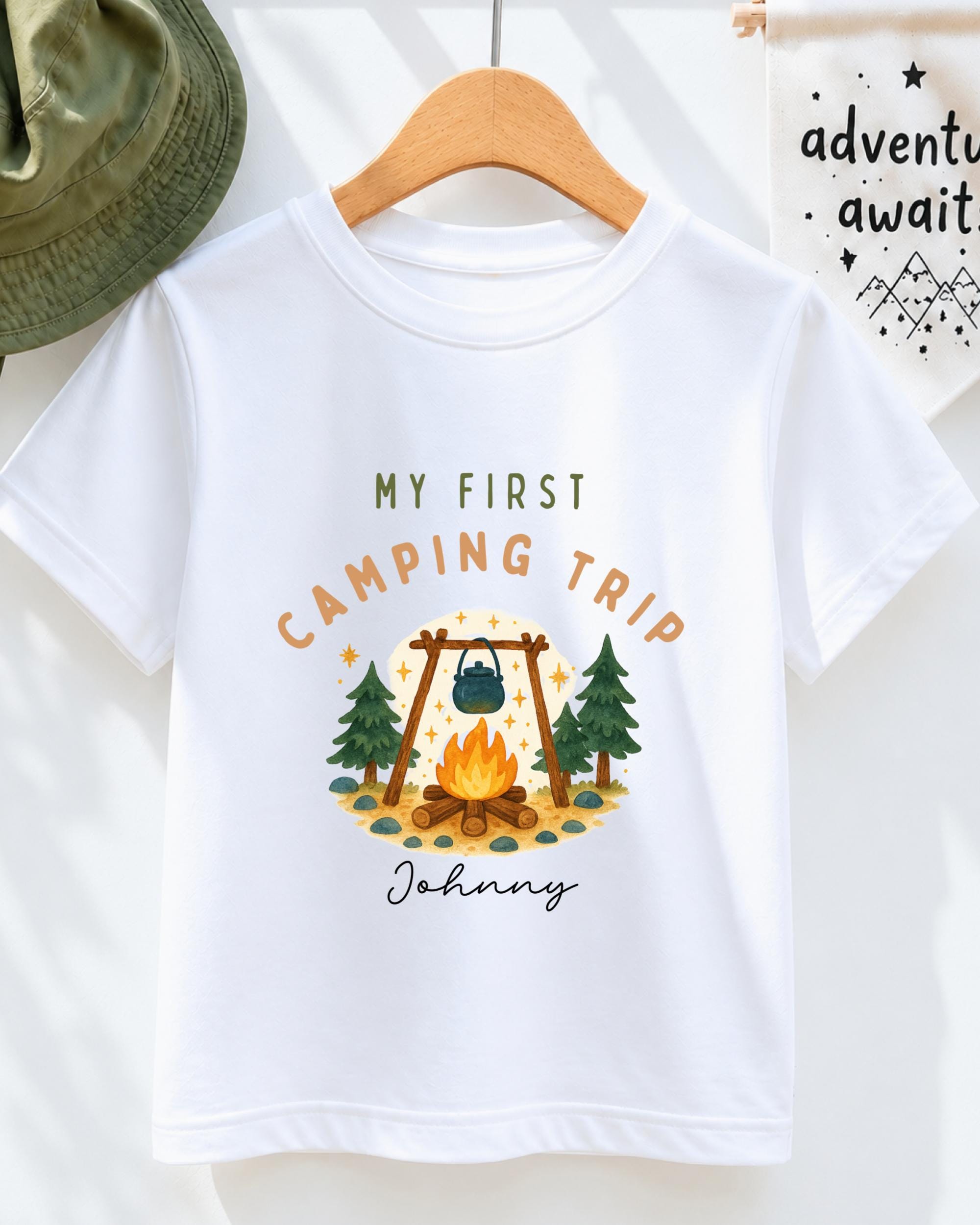 A white t-shirt with a printed design depicting a camping scene, including a campfire, pine trees, and mountains. The text on the t-shirt reads "My first camping trip" and "Johny".