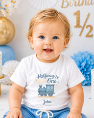 A smiling baby boy wearing a white t-shirt with a train graphic and the text "Halfway to One" is sitting in front of a birthday party backdrop with gold and blue decorations.