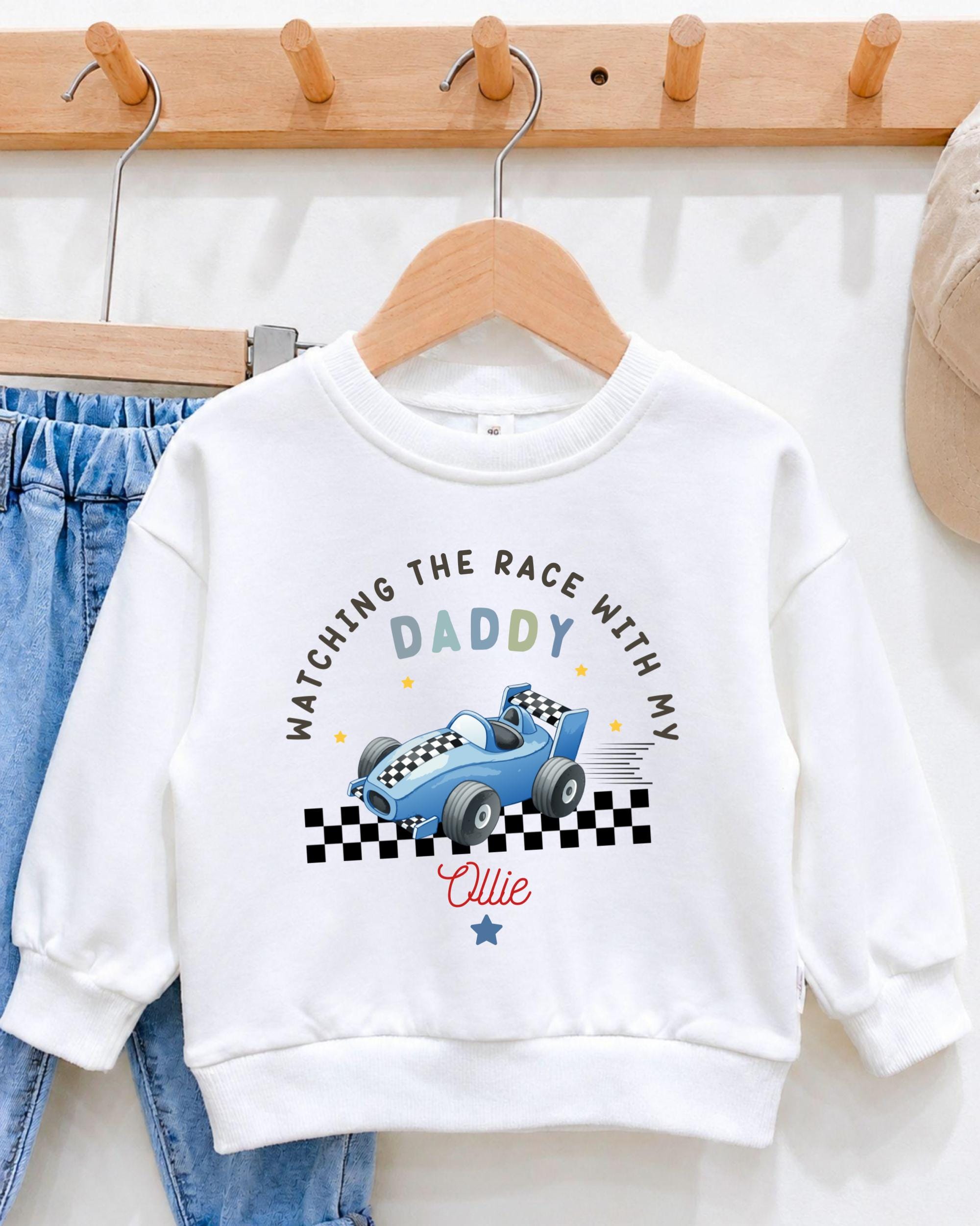 A white sweatshirt with a racing car design and the text "Watching the race with my Daddy" printed on it, hanging on a wooden hanger.