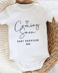 A white baby onesie with the text "Coming Soon" and "Baby Harrison 2026" printed on it, placed on a woven basket.