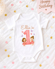 A white onesie with a pink number 1 and two fairy characters printed on it, placed on a bed with confetti and a "Happy Birthday" sign.
