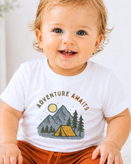 A smiling baby wearing a white t-shirt with a graphic design of a tent and mountains.