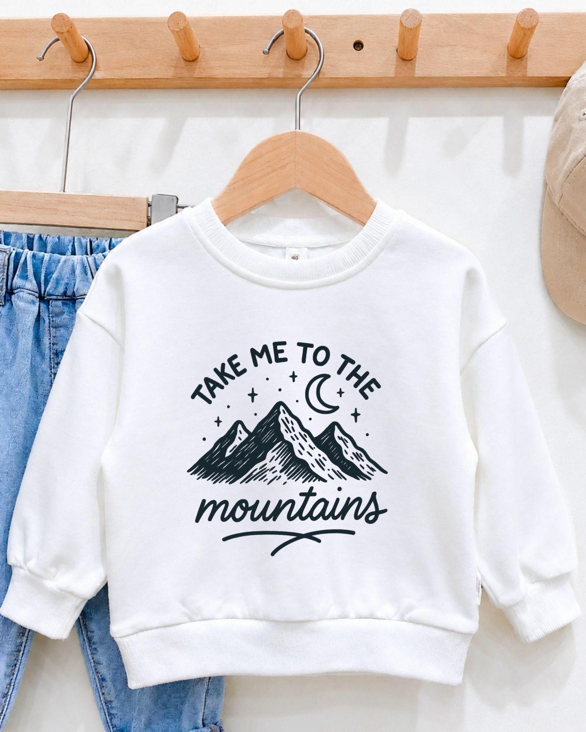 A white sweatshirt with a graphic design of mountains and stars, along with the text "Take me to the mountains" printed on it, hanging on a wooden hanger.