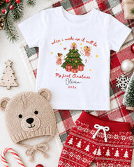 A white t-shirt with a Christmas-themed design, a beige knit hat, red and white patterned shorts, and other Christmas-themed items are arranged on a plaid blanket.