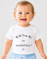 A smiling baby wearing a white t-shirt with the text "Will You Be my Godfather?" printed on it.