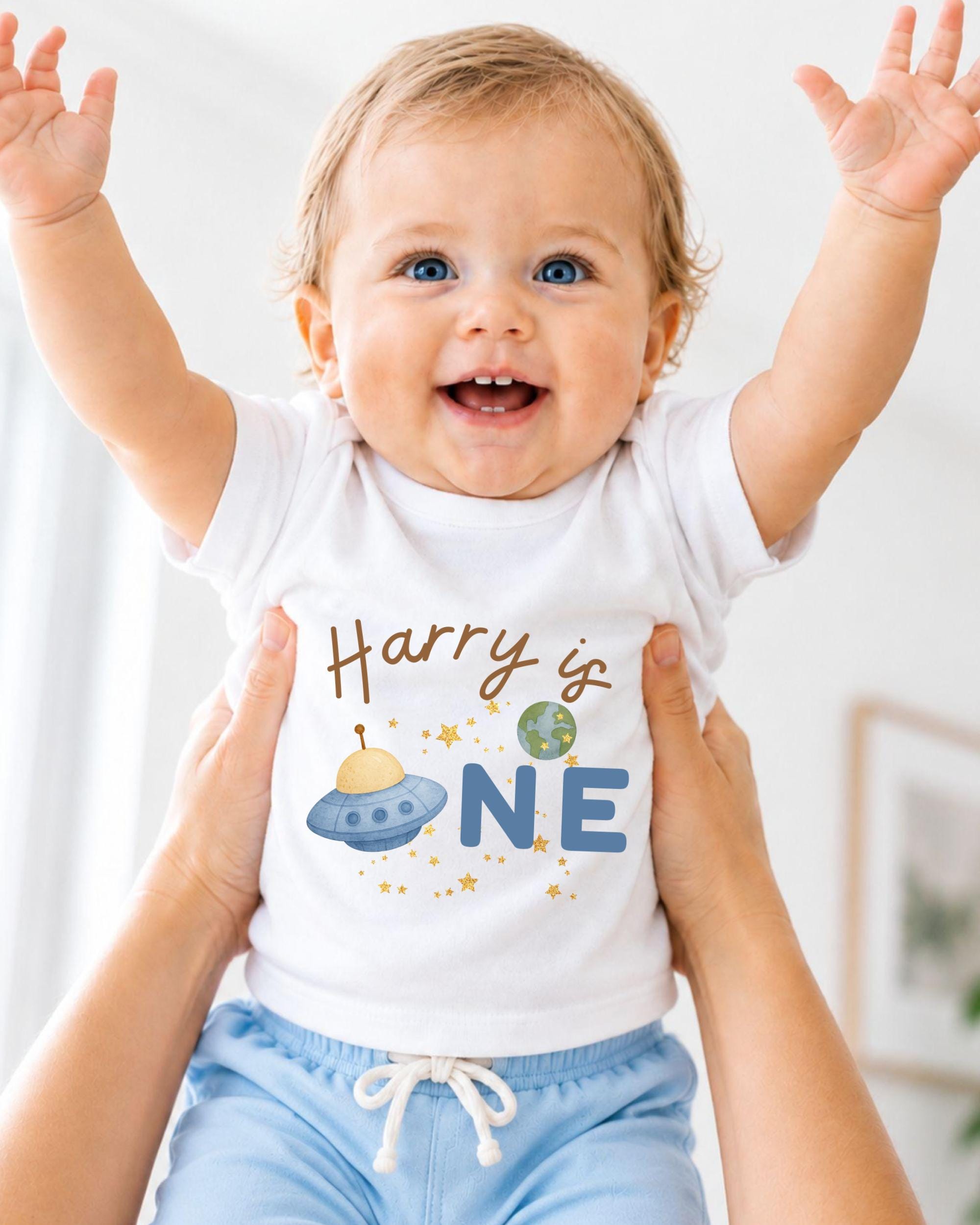 A smiling baby wearing a white shirt with a personalized text and an illustration of a spaceship.