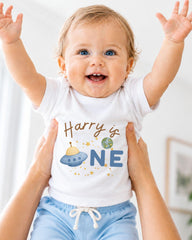 A smiling baby wearing a white shirt with a personalized text and an illustration of a spaceship.