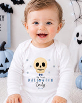 A smiling baby boy wearing a white onesie with a skull and snowflake design, sitting in front of Halloween decorations.