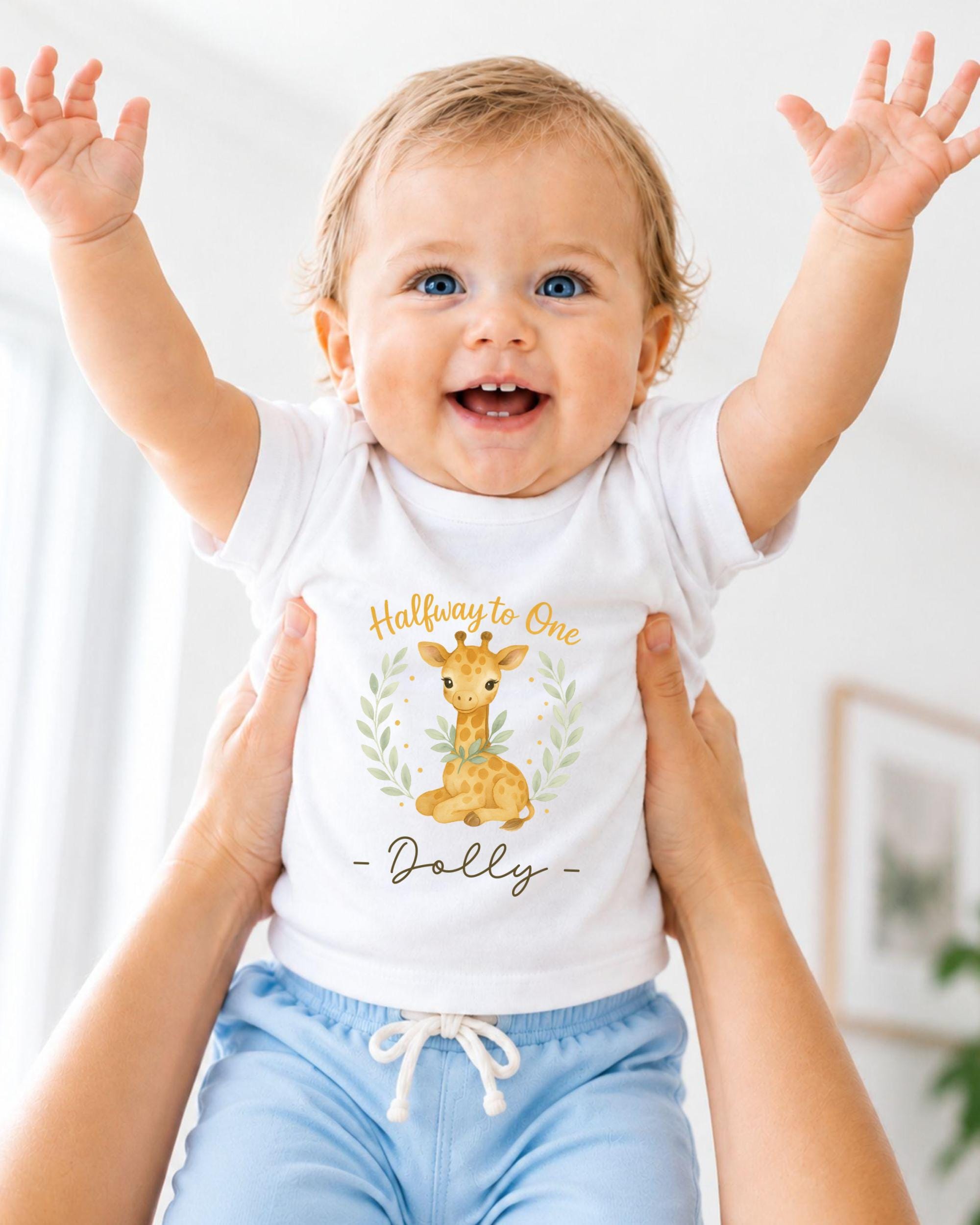 A smiling baby wearing a white t-shirt with a giraffe graphic and the text "Halfway to One Dolly" is being held up by someone's arms.