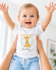 A smiling baby wearing a white t-shirt with a giraffe graphic and the text "Halfway to One Dolly" is being held up by someone's arms.