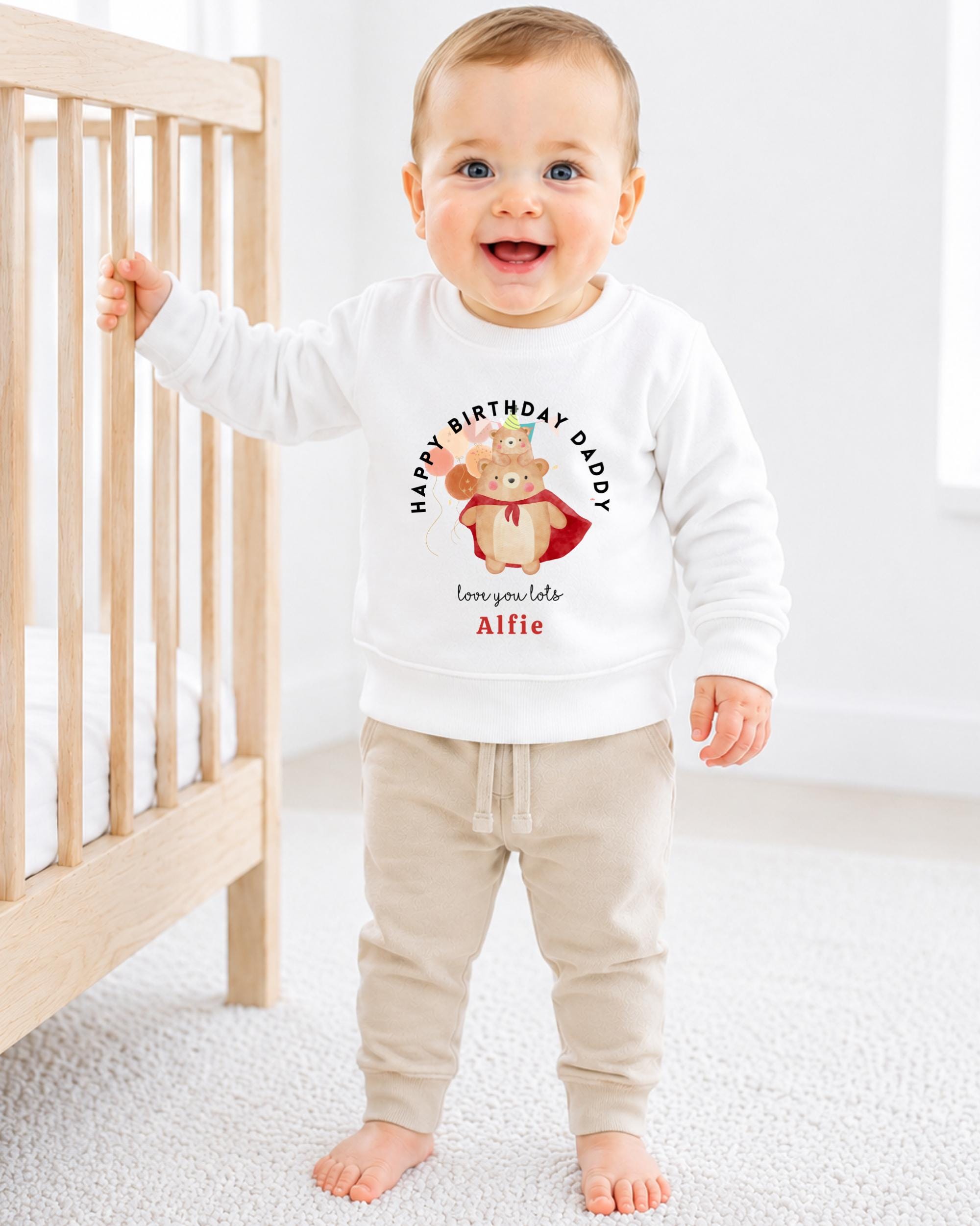 A smiling baby boy wearing a white sweatshirt with a cartoon bear and the text "Happy Birthday Daddy Alfie" printed on it.