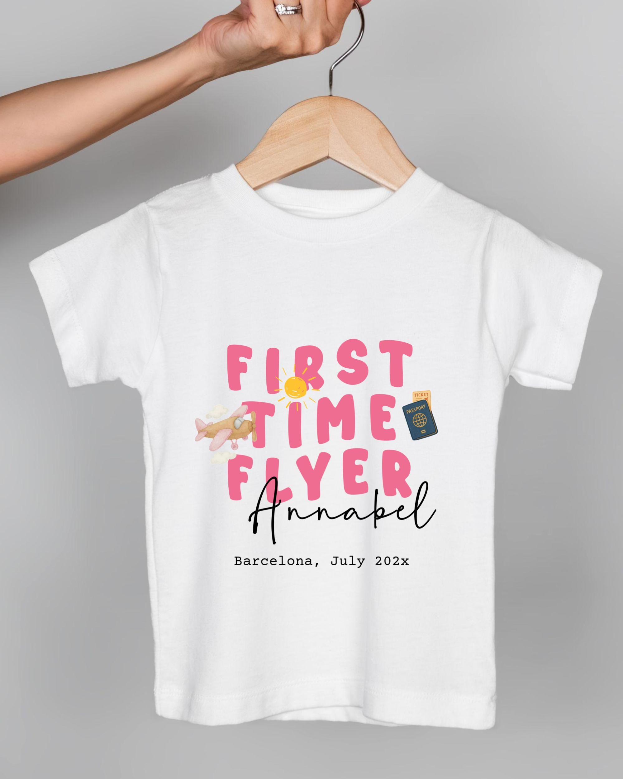 A white t-shirt with pink text and an illustration of an airplane, along with the text "FIRST TIME FLYER Annabel Barcelona, July 202x".