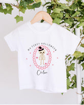 A white t-shirt with a Halloween-themed design hanging on a wooden hanger. The design features a skeleton wearing a witch's hat and surrounded by pink hearts.