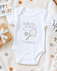 A white onesie with a tennis racket and stars printed on it, along with a "Halfway to One" sign and a gift box, all placed on a white blanket with confetti.